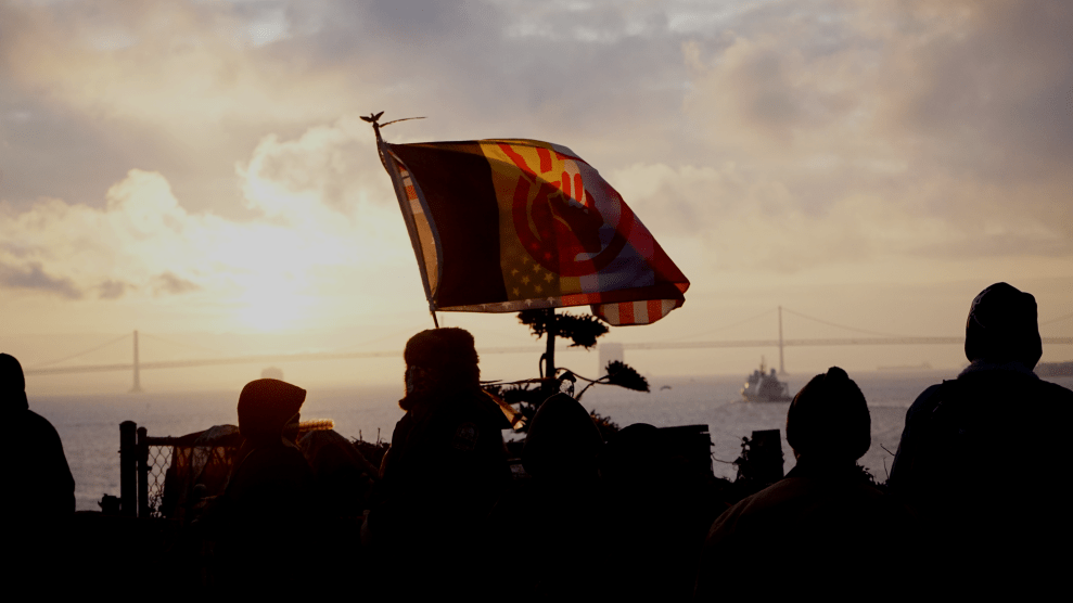 The American Indian Movement flag flies with an upside down American flag with the sunrise and SF Bay Bridge in the background.