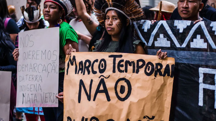 Three indigenous Brazilians dressed in some traditional clothing hold signs protesting the theory of marco temporal.