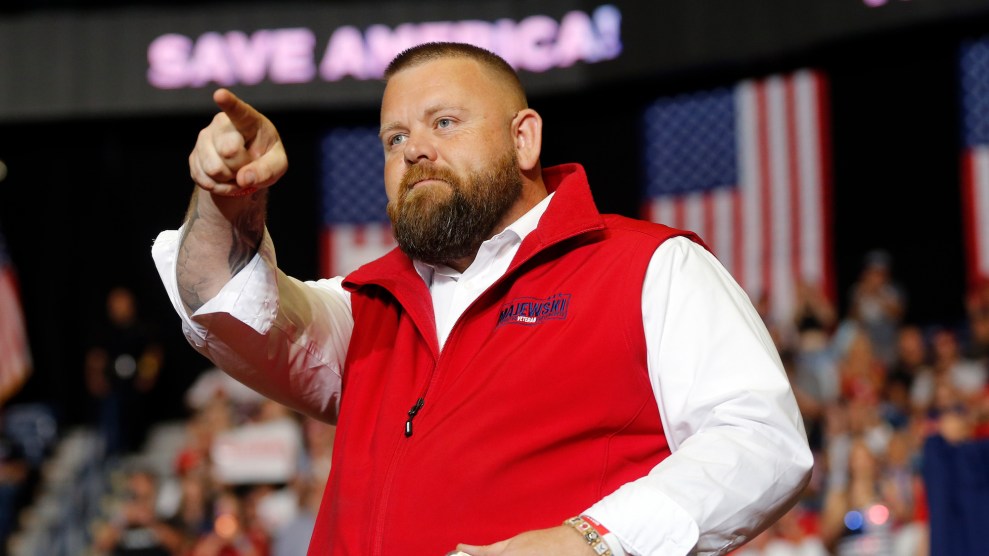 J.R. Majewski, a heavier white man with a beard, in a white shirt and red vest pointing to something off stage with a crowd surrounding him and a sign saying "Save America" in the background
