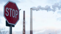A stop sign next to a Shell refinery, which smoke can be seen coming out of