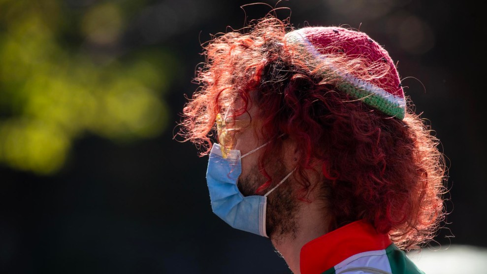 A person with pale skin and red curly hair looking to the side, wearing a blue surgical mask and a watermelon kippah.