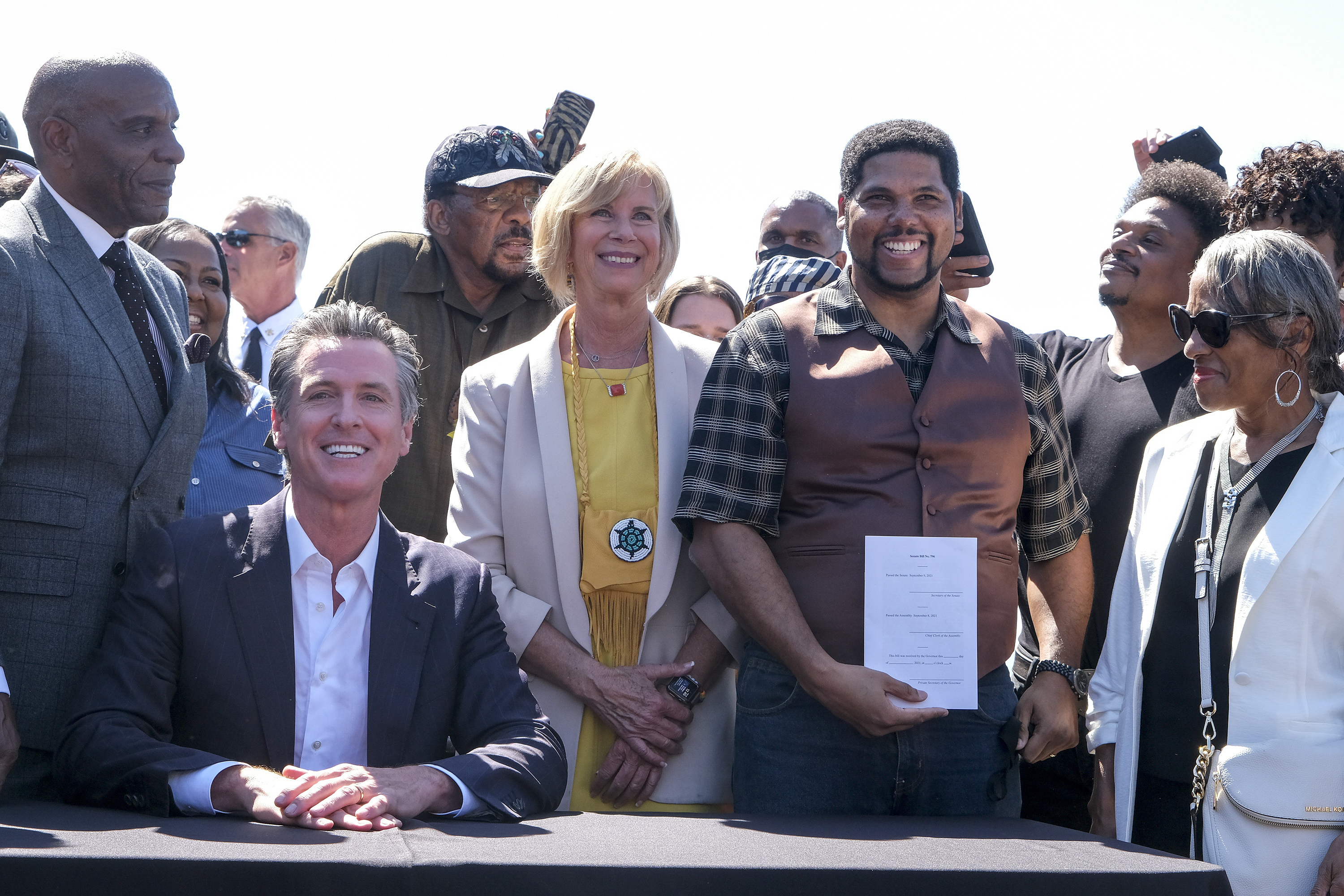 Group of people standing around California Governor Gavin Newsom, who is seated, smiling. Man holding a white document next to Newsom.