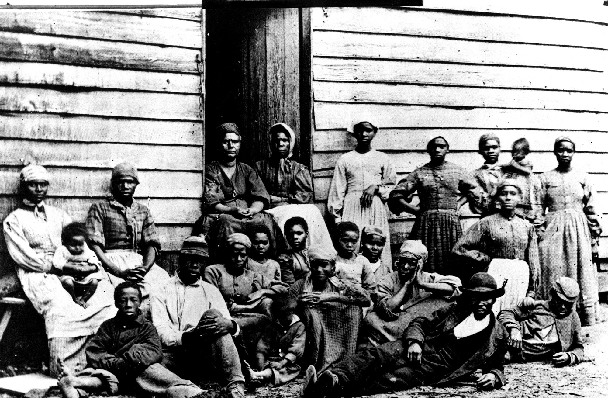 Black-and-white photo of a group of people sitting and standing in front of a wood building.