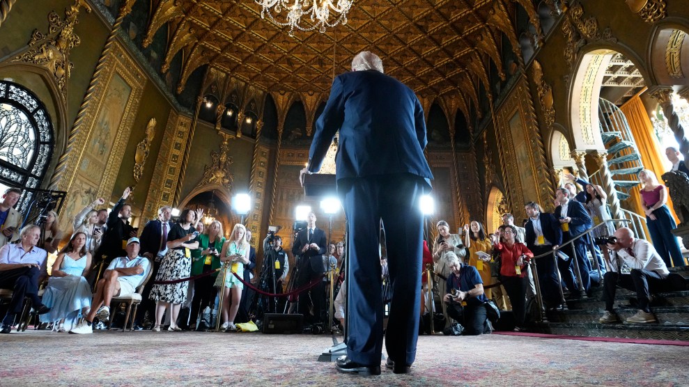 A full-length back view of Donald Trump standing at a lecturn, facing a row of reporters in a gaudy, gilded room.