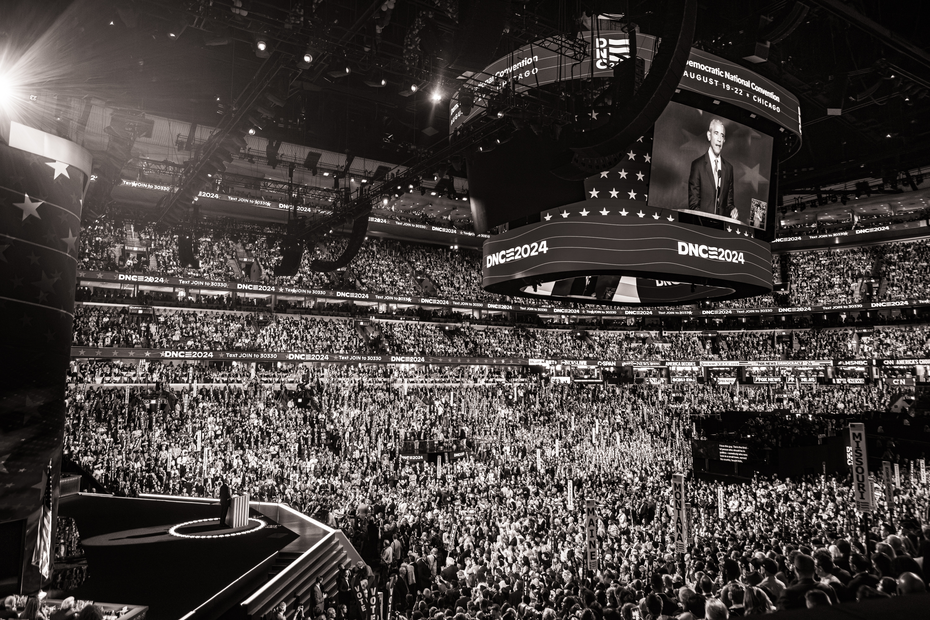 Black and white photo of the large crowd at the United Center for the DNC. Video image of Barack Obama overhead.