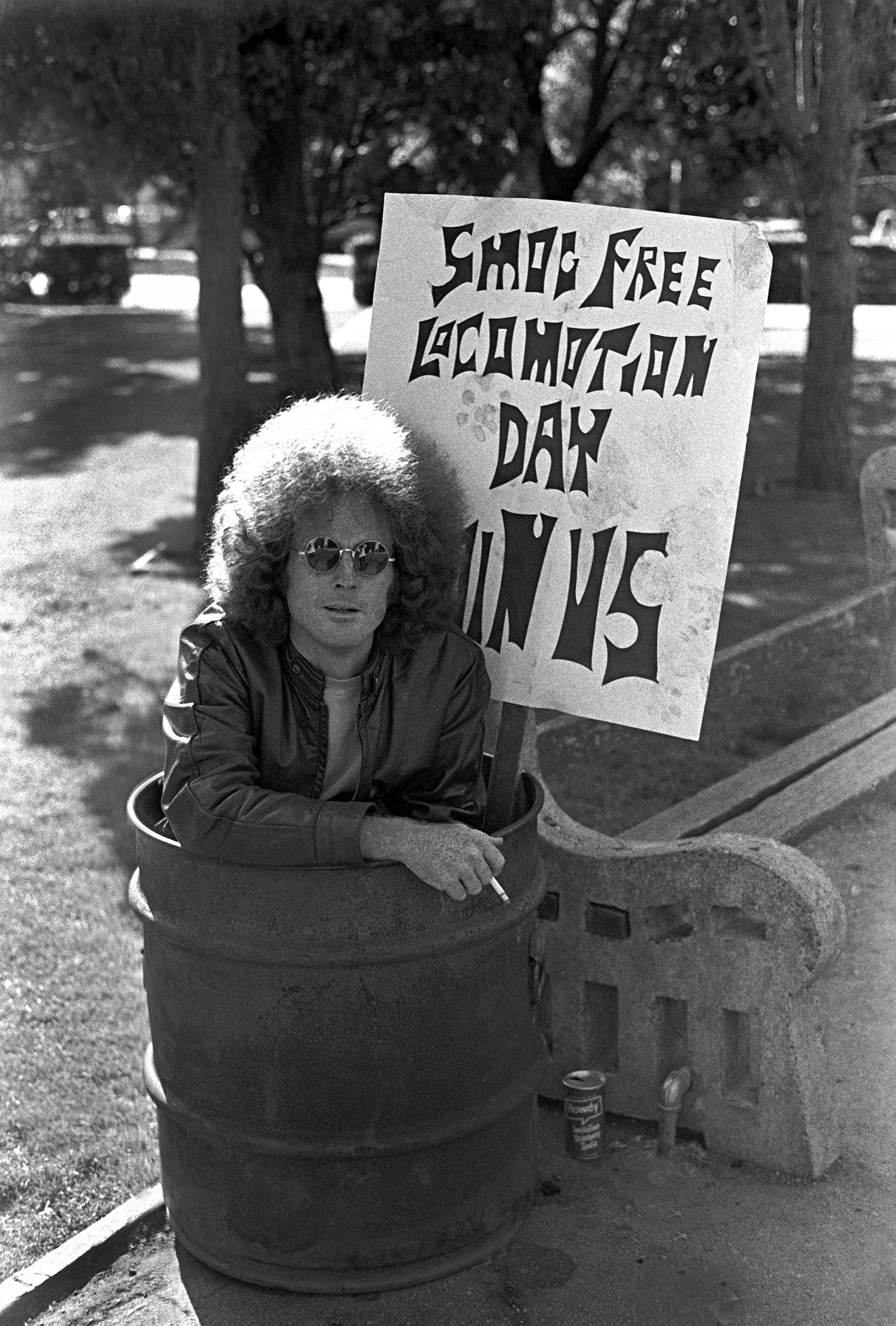 Black and white photo of a man with sitting in a trash can with a cigarette and a sign that reads, "Smog Free Liberation Day Join Us."