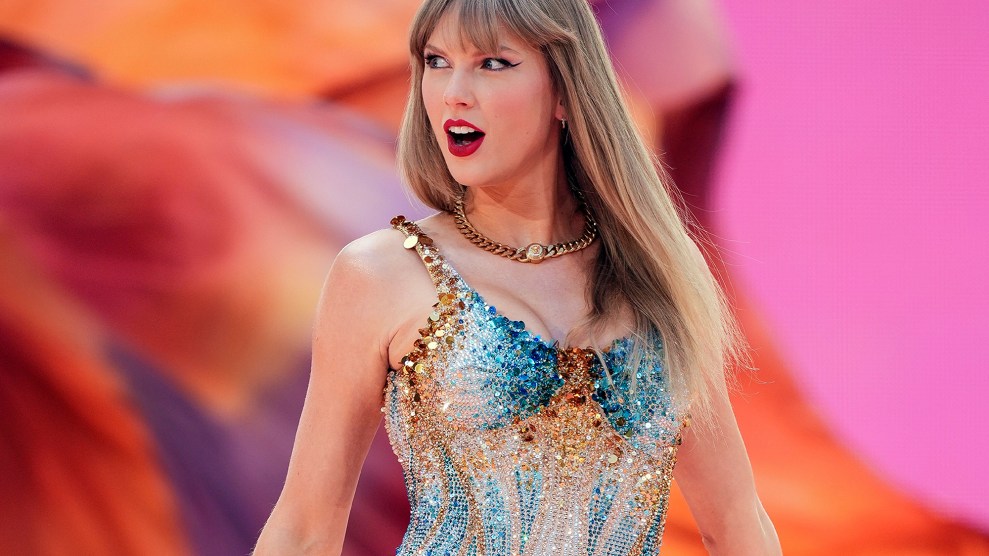 Taylor Swift, standing on stage during a performance. She is wearing a sparkly, fitted bodysuit with shimmering blue and gold embellishments.