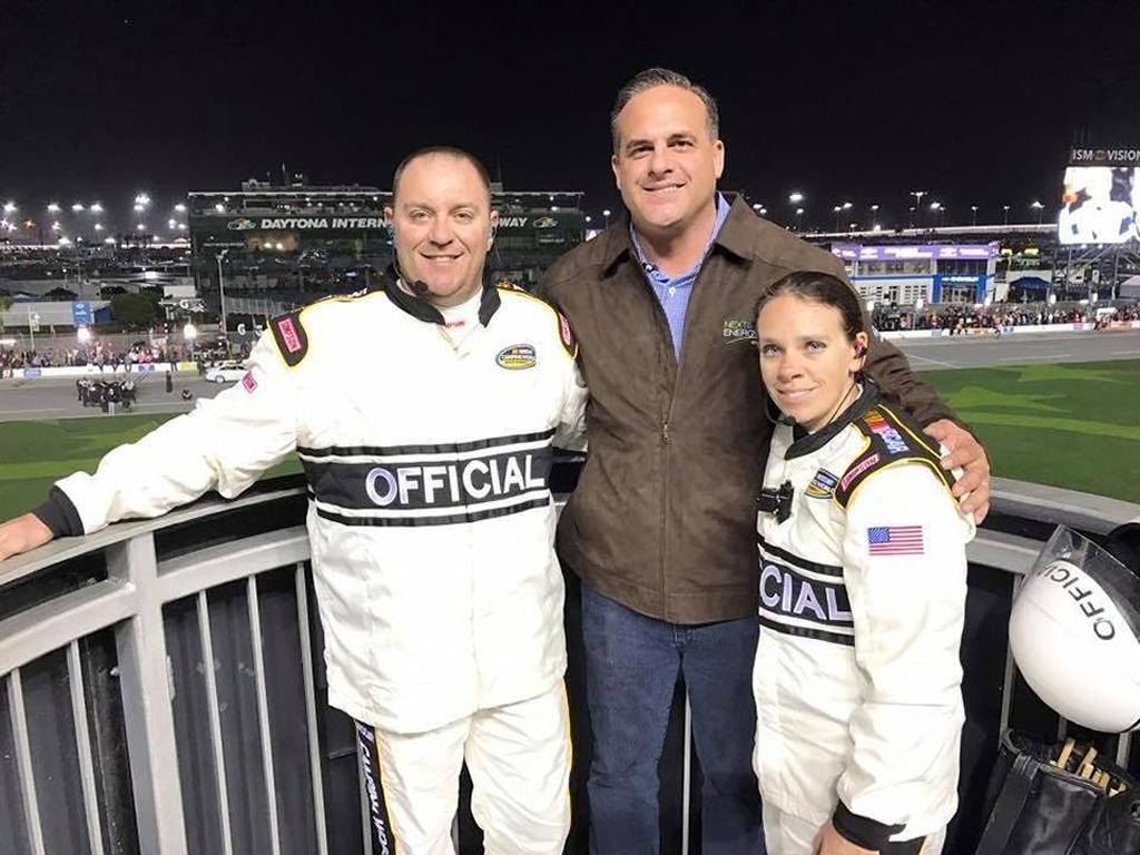 Man in brown jacket standing in the middle of a man and woman in white race car driving suits.