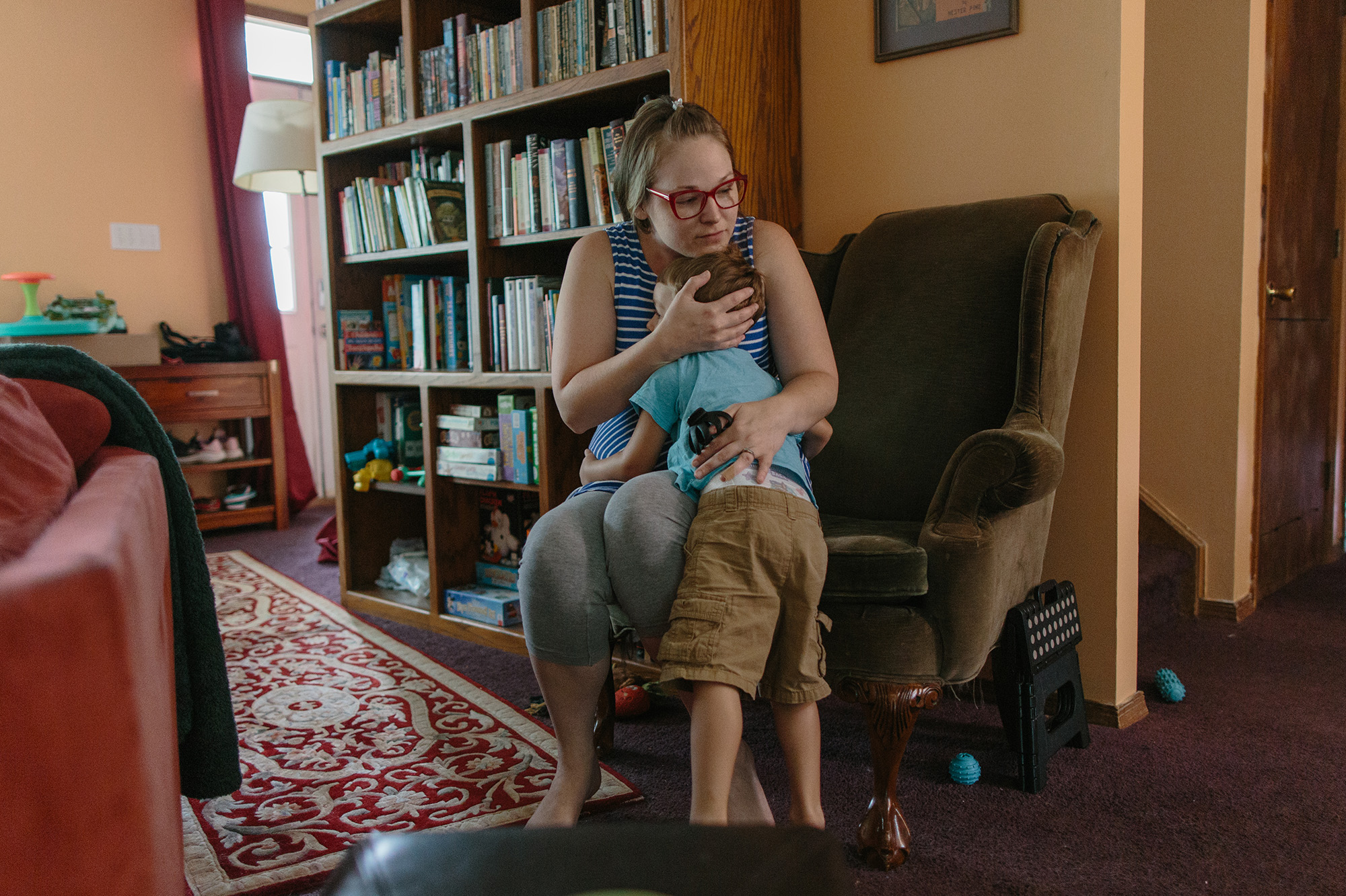 Grace Smith, wearing glasses, a blue-and-white striped tank top, and gray leggings, hugs her child Julian while sitting on a sofa chair.