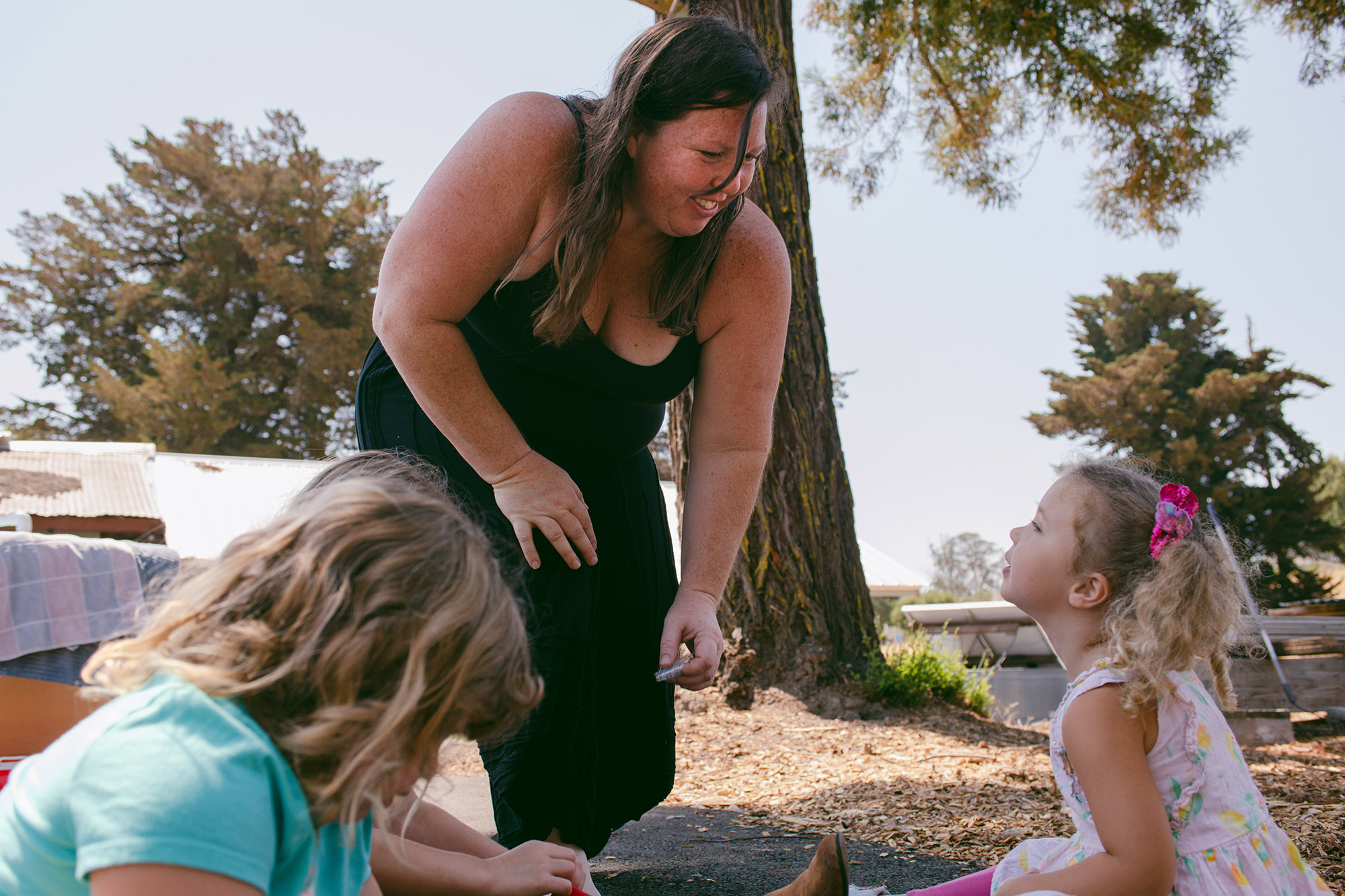 Susan Horton, wearing a black tank top and black pants, leans while she stands to talk to one of her daughters. In the foreground, another daughter, wearing a light blue shirt, is looking at the floor.