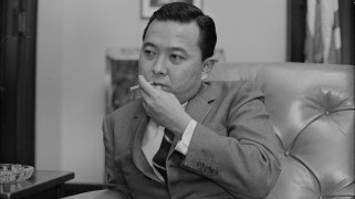 Senator Daniel Inouye, a man of Japanese descent, sitting on a chair and a suit and smoking a cigarette. His suit is loose near the area where his amputated arm would be.