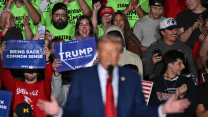 A photo of out-of-focus Donald Trump standing behind the podium, speaking to the crowd at a rally. Behind him, in focus, are people with pro-Trump signs, wildly cheering.