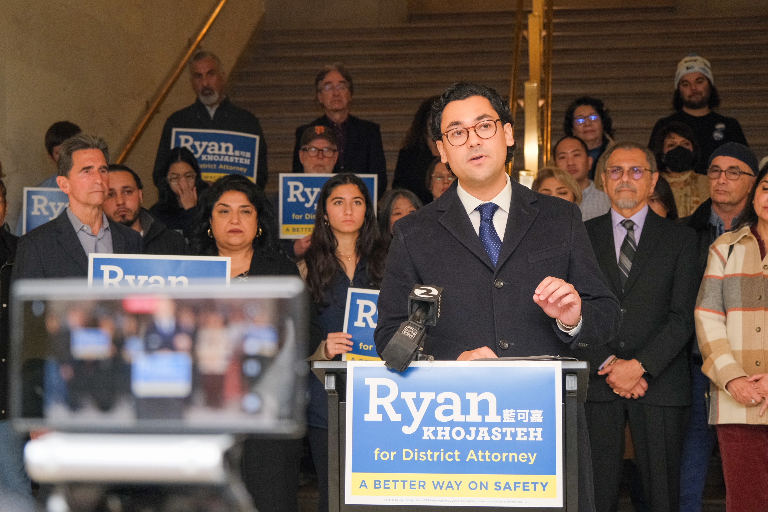 Ryan Khojasteh, wearing glasses along with suit and tie, gives a speech behind a podium. On the front of the podium is a campaign sign that reads: Ryan Khojasteh for District Attorney: A Better Way on Safety. About 20 people stand behind him in support of his candidacy. 