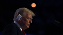 Donald Trump, seen in profile from his right in front of a bank of microphones, tilts his head downward with eyes closed. He has a bandage on his right ear.