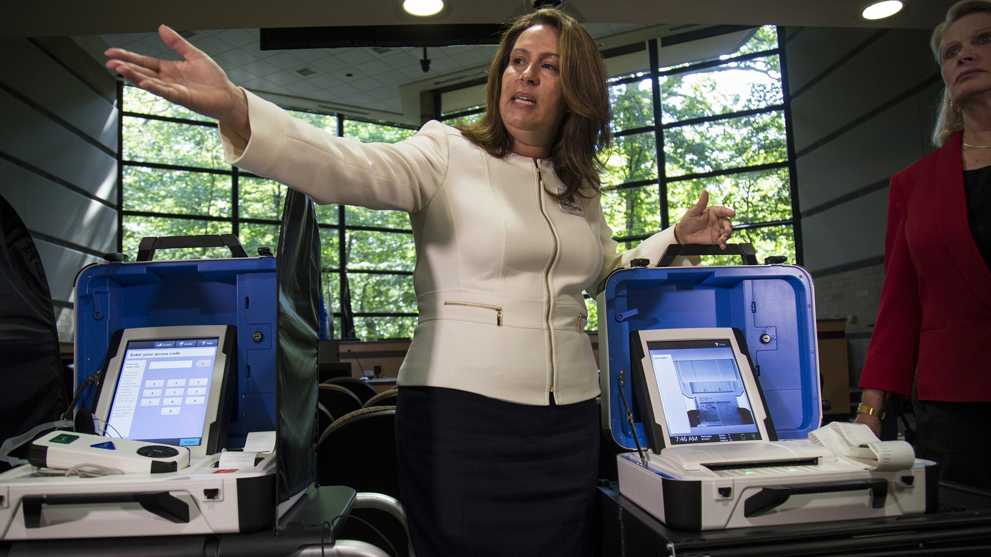 Woman in white stands in front of two pieces of voting equipment, gesturing with an outstretched hand.