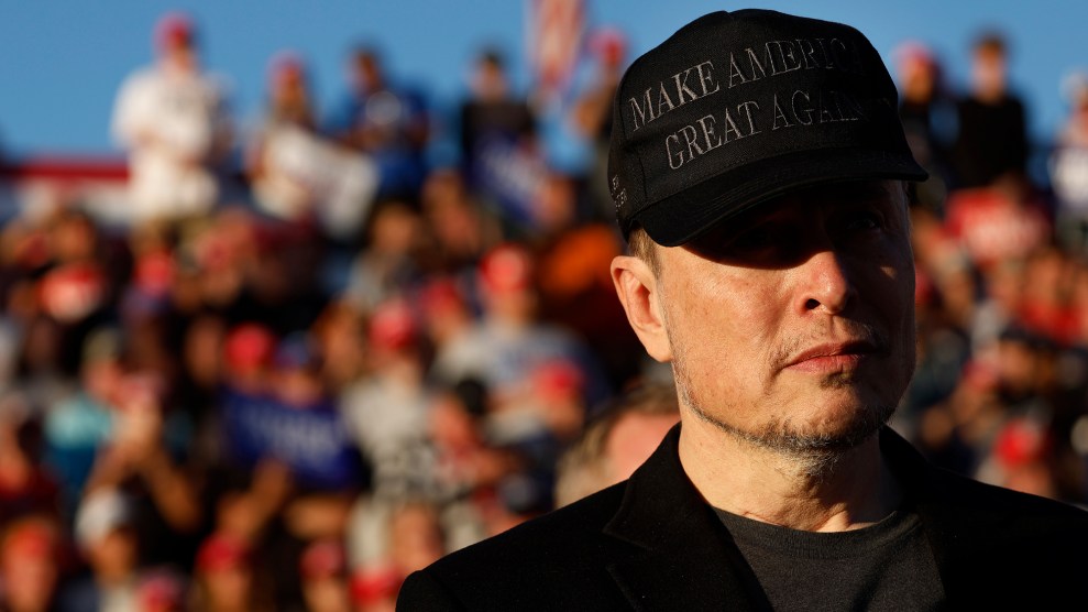 Elon Musk wears a black "Make America Great Again" ball cap while attending an outdoor campaign rally for Republican presidential nominee Donald Trump. The bill of the cap casts a shadow over Musk's eyes. The sport jacket and t-shirt he is wearing are also black, though the t-shirt is a slightly lighter shade. Behind Musk, out of focus, are bleachers full of Trump supporters.