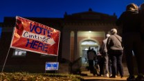 A photo of a dozen voters stand on a walkway outside the Cincinnati Observatory, which is a Greek Revival style building. The people are lit up by a street lamp as they've arrived in the early morning hours. In the grass beside the walkway are two signs. The red sign with blue type reads: Vote Here: Cinti 5E and it has a white arrow pointing toward the door. A blue sign in the distance has a white arrow that points the opposite way and reads "Handicapped Access."