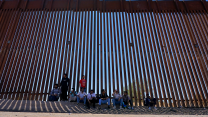 Ten people sit in front of a tall border fence.