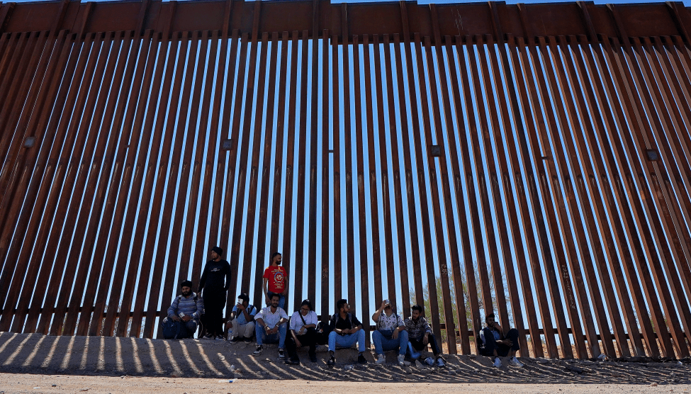 Ten people sit in front of a tall border fence.