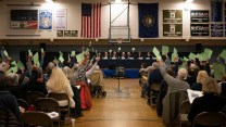 School gymnasium filled with people sitting in folding chairs, some with their arms raised, holding pieces of green paper.