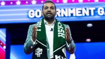 Kash Patel, wearing a green jacket and scrarf reading "fight with Kash," speaking at CPAC