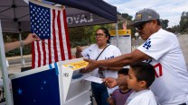 A Hispanic man and woman stand with their two children around a ballot box. The man and woman wear white Los Angeles Dodgers jerseys. Their two young children wear t-shirts. The polling station is located outdoors, near a portable tent. From the edge of the tent hangs an American flag. A poll worker's arm reaches into the photo, in an attempt to hand a pen to the woman.