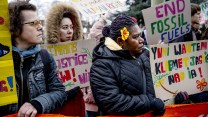 Three people hold rainbow signs in different languages. They are bundled in dark jackets and have serious looks on their faces.