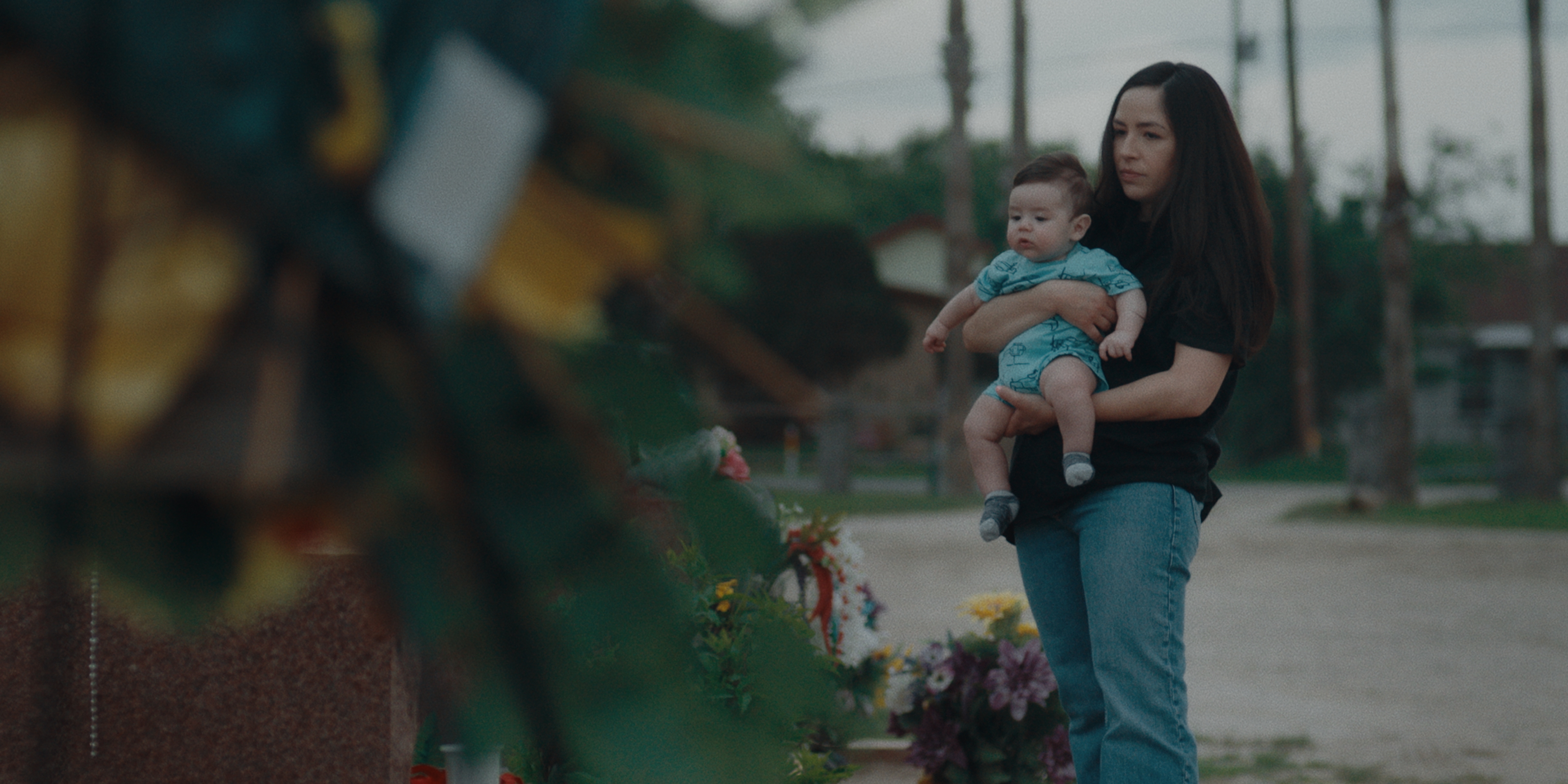 A still image from a documentary of a woman(Alejandra) standing at the grave site of her mother Melba while holing her child. Flowers are out of focus in front and behind the woman(Alejandra).