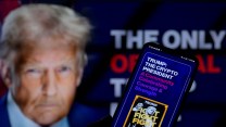 A cellphone in the foreground shows a screen that says, "Trump: The crypto president." Donald Trump's face is prominent but out of focus in the background.