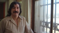 Portrait of a man with light brown skin, black hair, and a black mustache, standing next to a bolted window. He is wearing a light brown button down shirt.