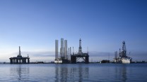 Oil rigs silhouetted black in the distance, mirrored into blue water. Blue-white sky in the background.