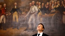 Elon Musk, in black suit with white shirt and black tie, looks up toward the ceiling of the US Capitol building with a wry smile. Behind him is the John Trumball painting “Surrender of General Burgoyne.”