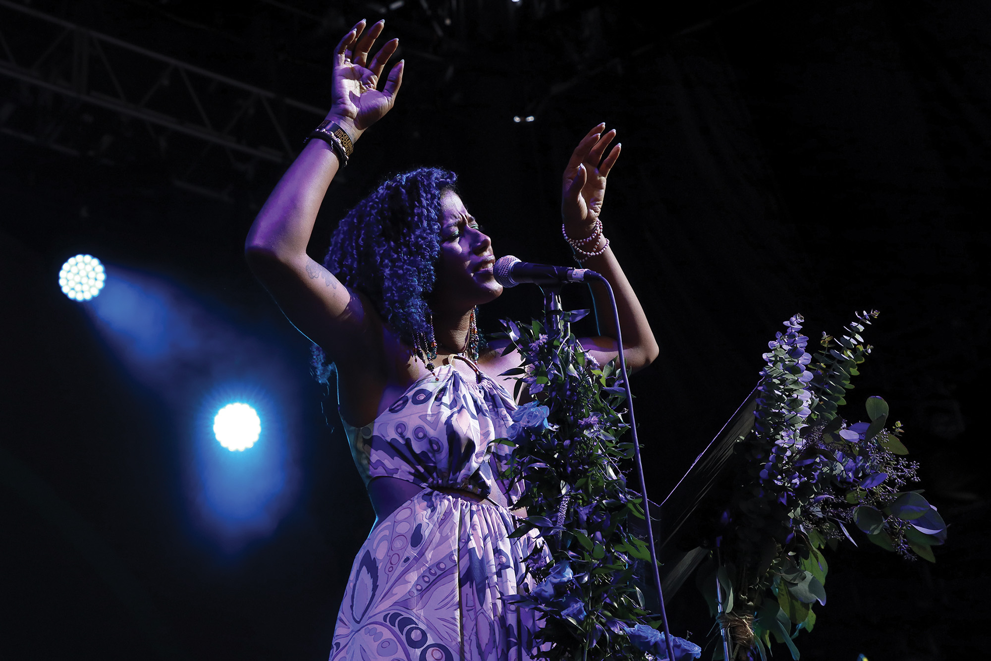 Aja Monet performing in front of a microphone with her eyes closed and hands raised.