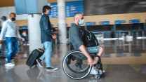 A white man in a wheelchair wearing a surgical mask rolling through an airport