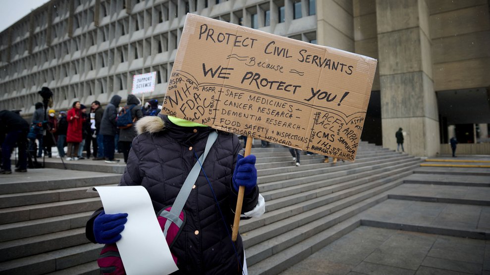 A person standing in front of a large brutalist building, wearing a navy blue coat and royal blue gloves, holds up a cardboard sign on a pole in front of their face. The sign reads in part, "Protect Civil Servants Because We Protect You!" In the background, many more people in colorful coats, standing on the raised steps in front of the building, are gathered to protest DOGE, as well.