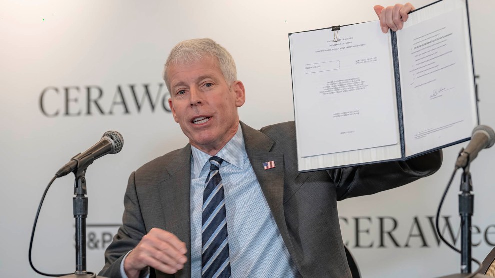 A white man wholes up a white paper. He sits in front of a microphone. He has white hair and a blue shirt.