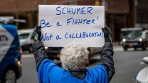The back of a person's head, white hair, blue shirt. Sign that says "SCHUMER be a fighter! Not a COLLABORATOR."