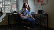 A White female doctor, wearing glasses, a gray jacket and blue scrubs, sits at her desk. Papers of children's hand drawings hang on the wall.
