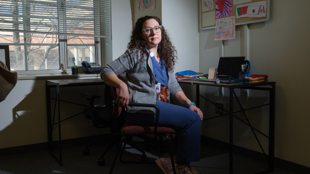 A White female doctor, wearing glasses, a gray jacket and blue scrubs, sits at her desk. Papers of children's hand drawings hang on the wall.
