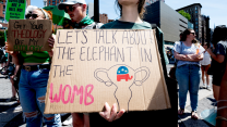 People hold up signs at an abortion protest. In the front is a young woman holding a brown cardboard sign that reads, "Let's talk about the elephant in the womb." At the bottom of the sign is an outline drawing of a womb with the GOP elephant logo inside.
