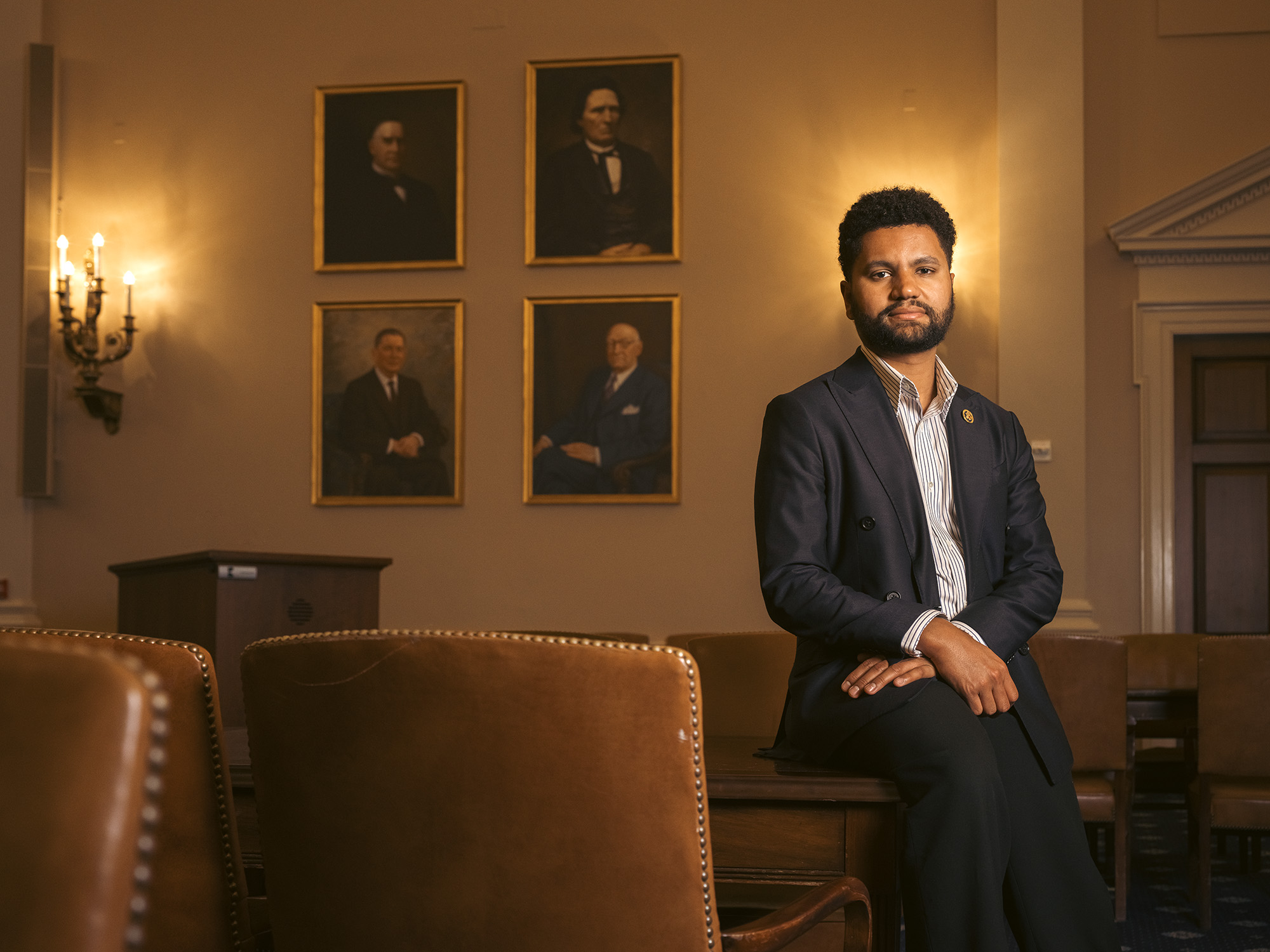Rep. Maxwell Frost sitting on a table in a room with four portraits of old white men on the wall.
