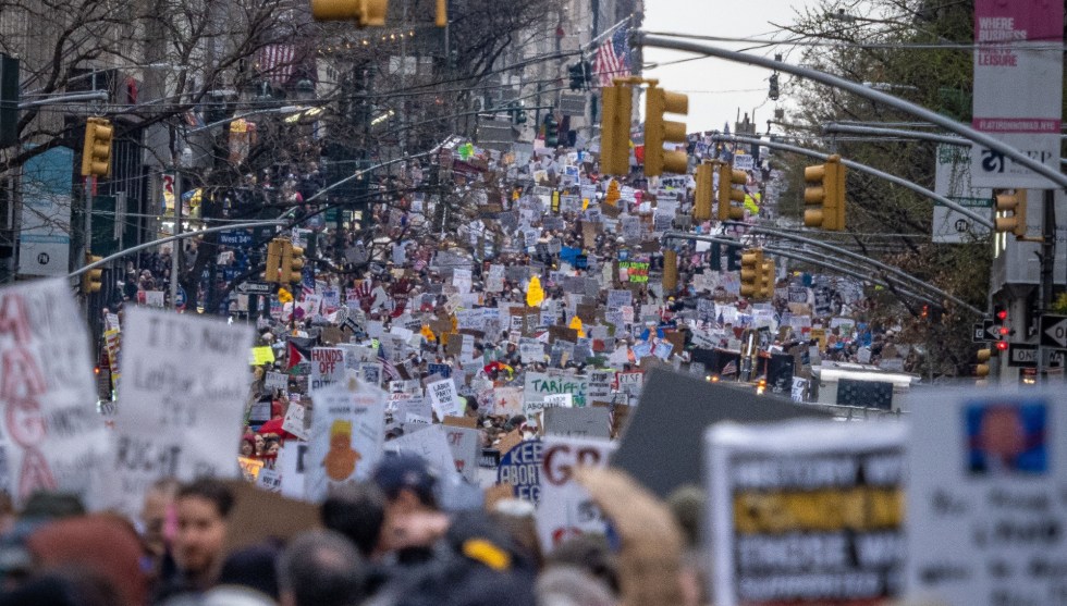 An endless crowd with many holding signs fills a city street, with street lights and lamps overhead.