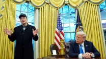 Donald Trump sits at a desk, looking over his right shoulder toward Elon Musk, who stands beside the desk.