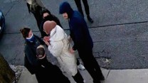 Seen from above in surveillance camera footage, a young woman in a white coat and pink hijab is detained by several masked plainclothes individuals.