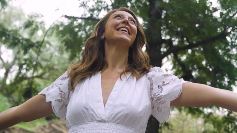 A woman with red hair stands in front of trees in a white sundress with arms outstretched.
