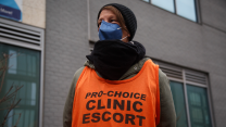A photo of a woman bundled up in a brown coat, black scarf and black beanie who is looking into the distance, as if on guard, as she stands outside an abortion clinic. She wears a blue face mask and an orange vest that reads in black sans serif letters "PRO-CHOICE CLINIC ESCORT."