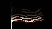 A close-up shot shows an American flag waving gently at night, barely illuminated by light from below. The light catches the fabric's folds, creating highlights and deep shadows, while the rest of the scene is enveloped in complete darkness. The flagpole is visible on the far left.