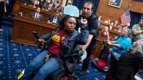 A Black woman with a fist above her head and a sign on her wheelchair saying :hands off our Medicaid" being rolled out by a US Capitol Police Officer. There are Congresspeople in the rows in the background,.