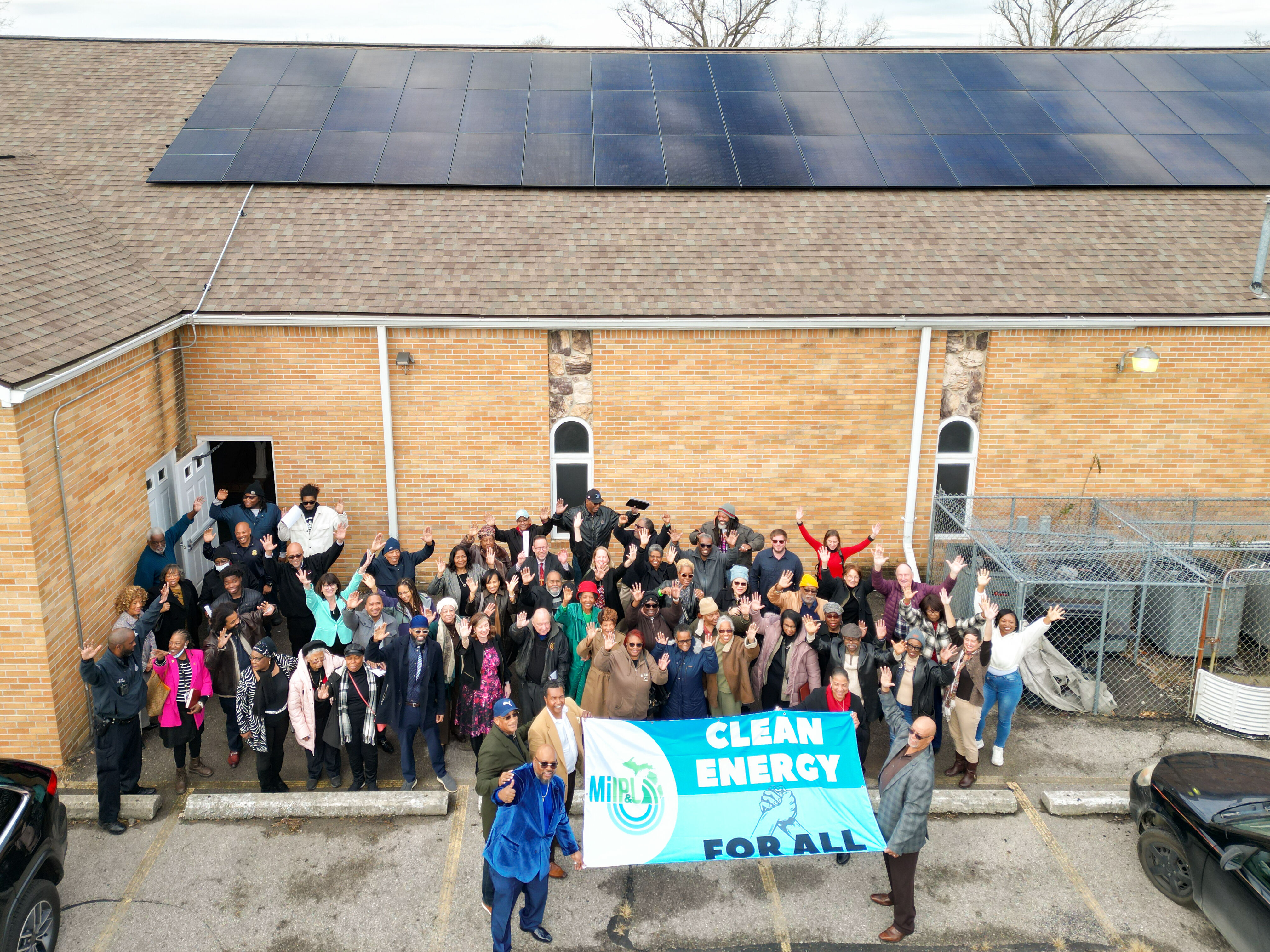 A group of people holds a sign that says "Clean Energy for Everyone" in front of a church with a solar panel