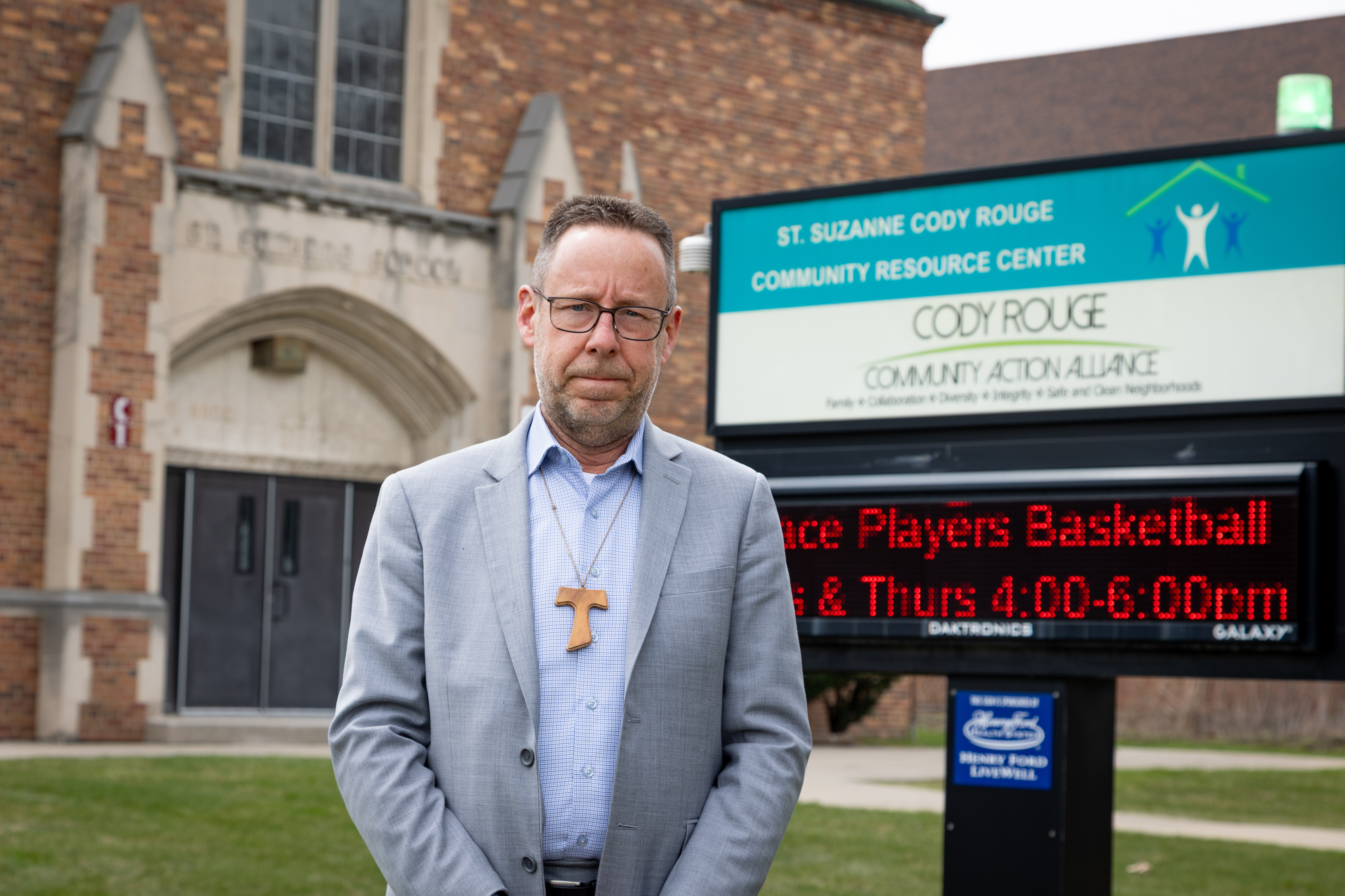 A white man wearing a cross stands in front of a church.